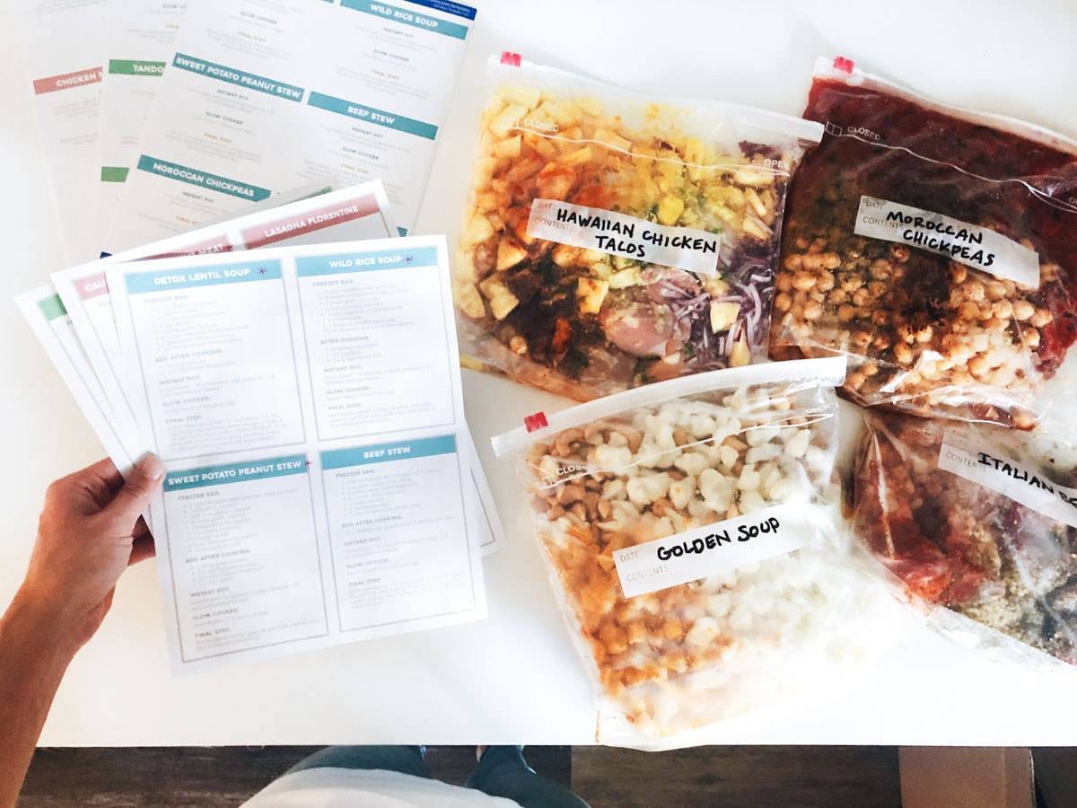 White hand holding meal templates for freezer meals. There are freezer meals on the counter in plastic bags.